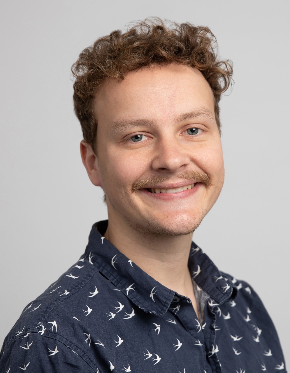 An image of Jayden Sercombe. He is a man with short dark hair, a brown moustache and is wearing a patterned black shirt. He is smiling in the photo. 