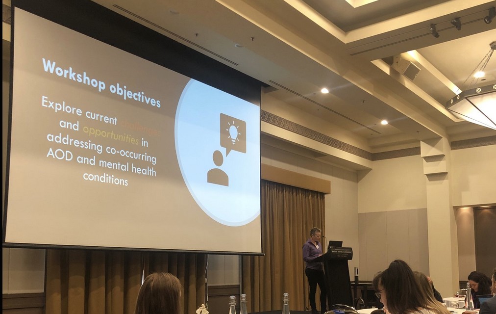A person standing in front of a podium with a large powerpoint in the background. 