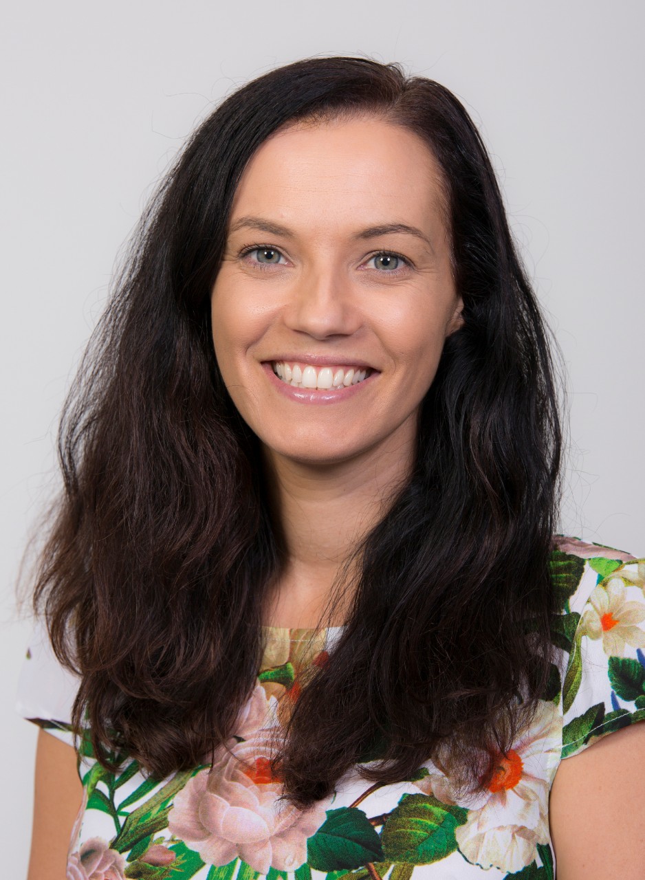 an image of Jo Cassar. She has shoulder length dark brown hair and is wearing a turqouise top. She is leaning forward and smiling. 