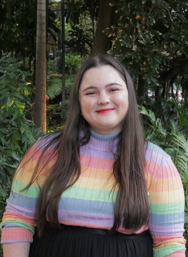 Grace has long, blonde-brown hair and is wearing a rainbow striped shirt with a denim vest over the top. 