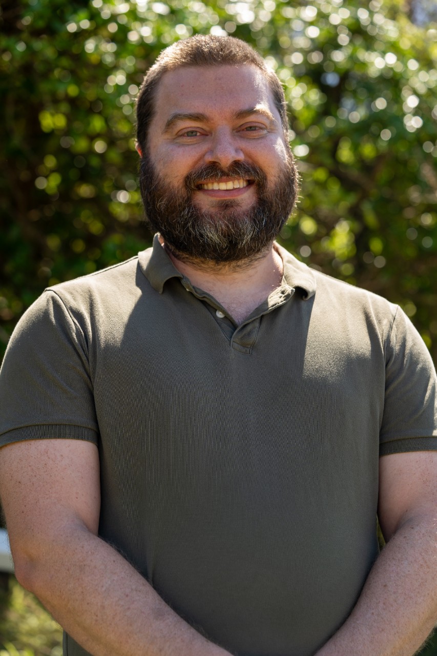 A man looks forward. He has short, curly brown hair and is wearing a dark green shirt. He is smiling. 
