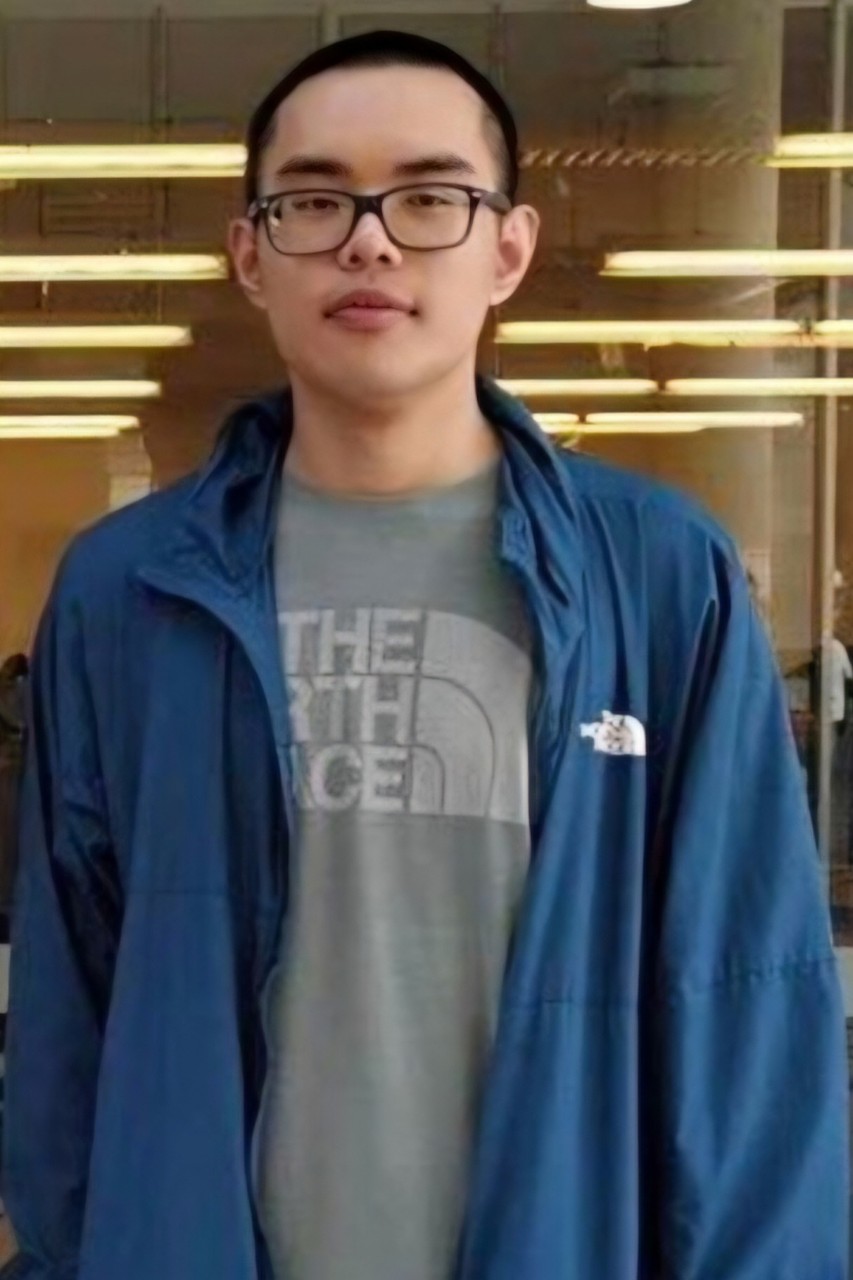 Tim is wearing a grey shirt with a blue jacket and has short dark hair. He is looking directly ahead.