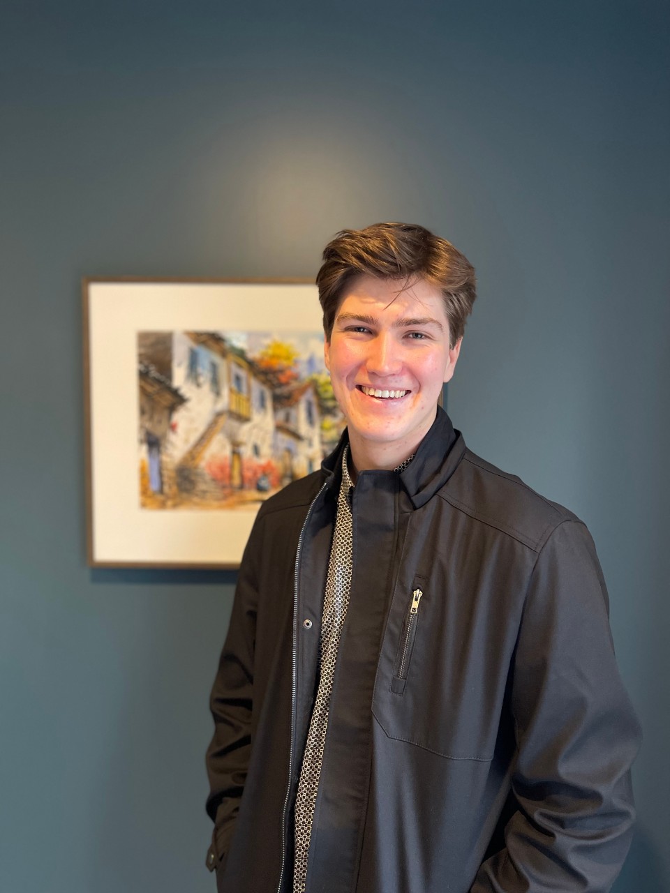 A headshot of Ben. Ben is wearing a brown shirt with a black jack on top, and has short, brown hair. Ben is smiling and standing in front of a landscape painting. 