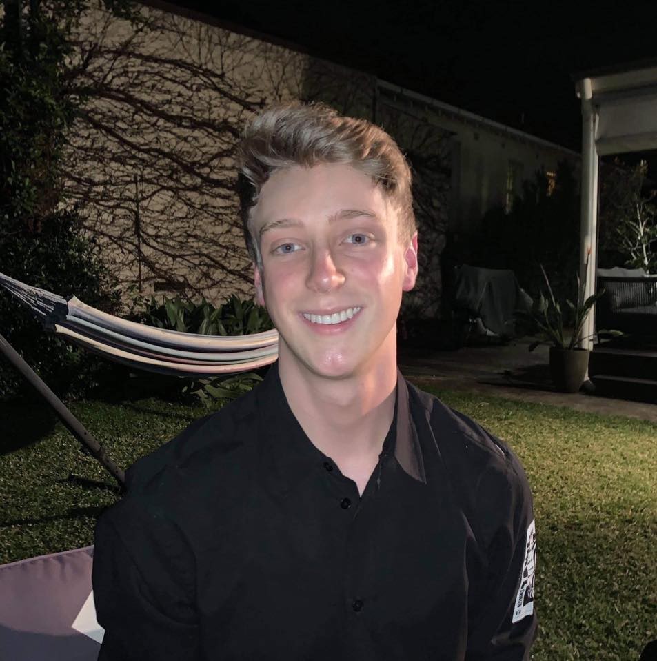 A headshot of Conor Hinds. He is a blond man who has short hair and is wearing a black shirt. 