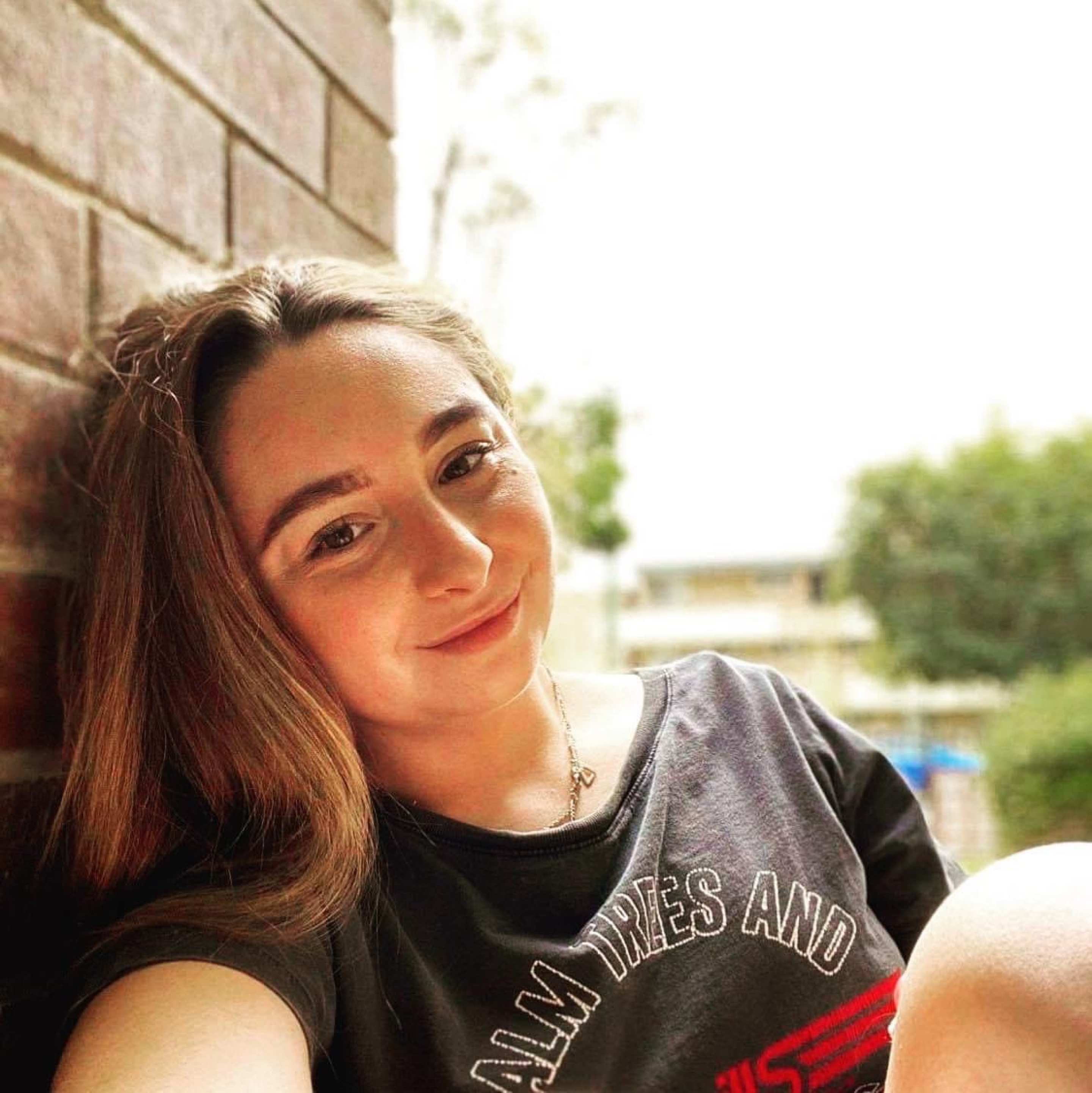 A headshot of Tanya. Tanya has long, brown hair and is wearing a black print t-shirt. Tanya is smiling.