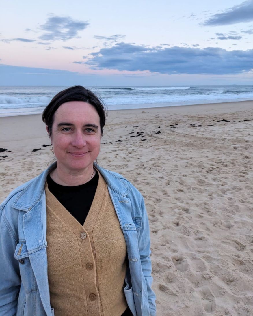 A photo of Jennifer on the beach wearing a brown cardigan and a denim jacket