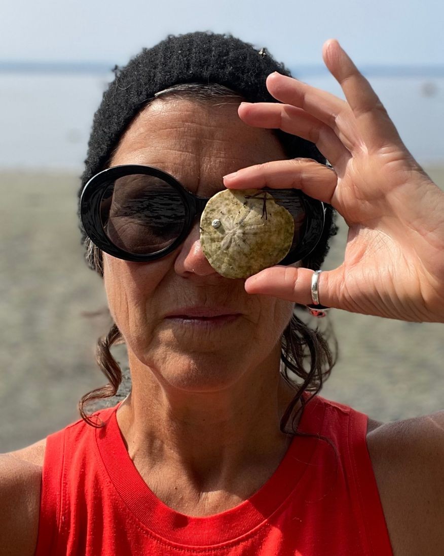 A photo of Astrida on a beach holding a sand dollar to her eye 
