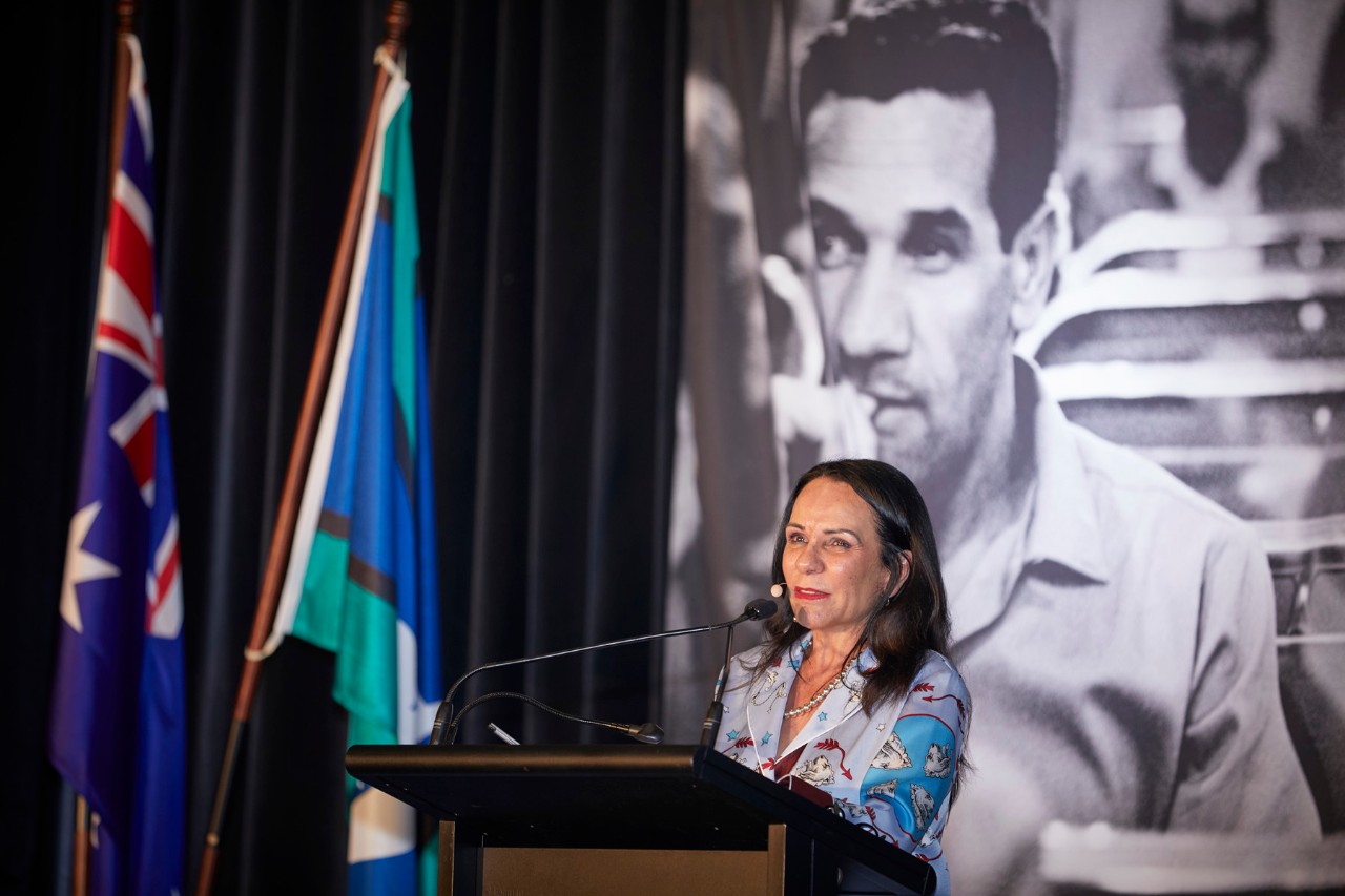 Linda Burney MP standing at a lecturn with a poster of Charles Perkins behind