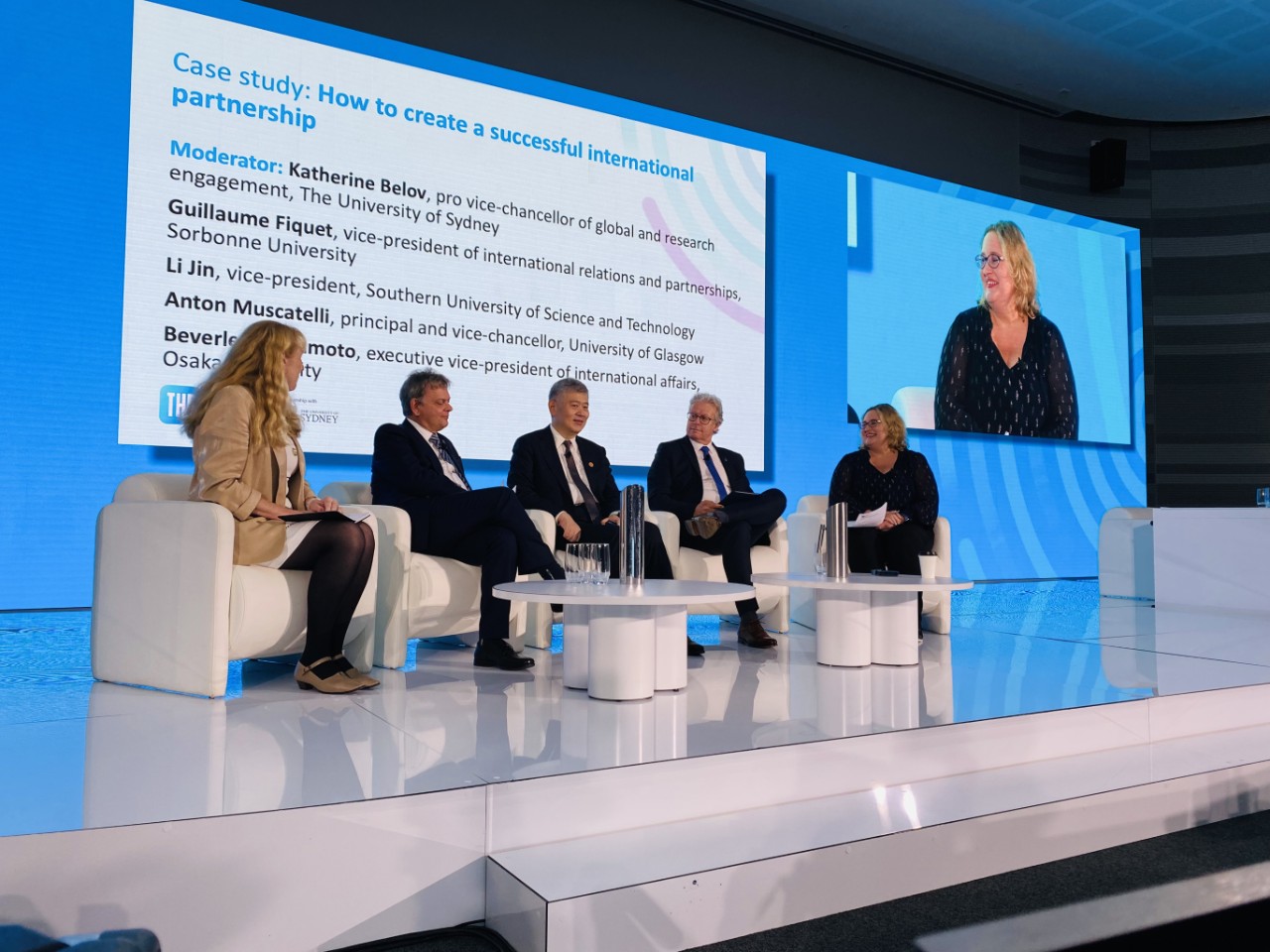 Five panellists seated on stage with screen displayed behind them