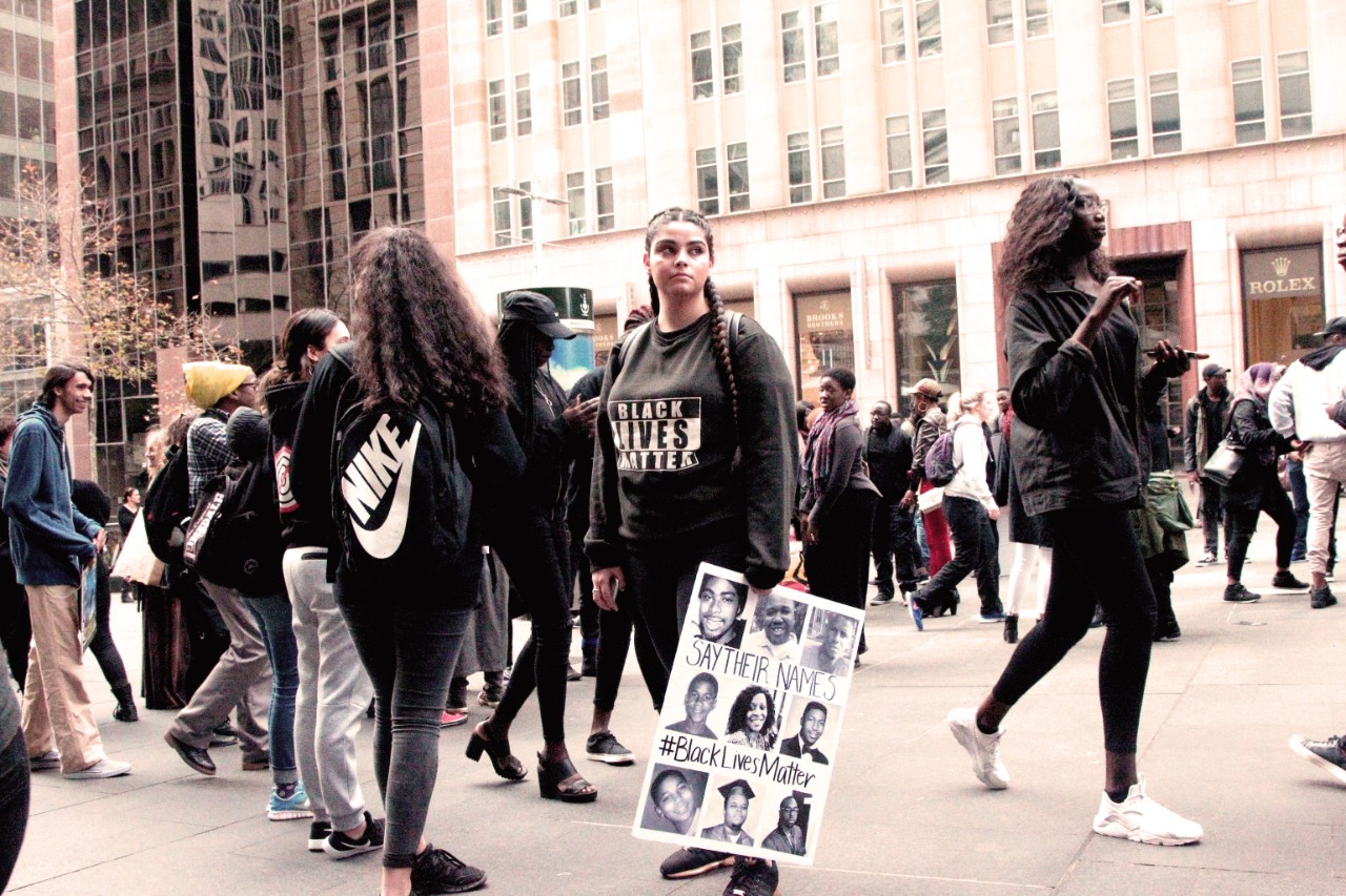 A photograph of three women at a black lives matter protest. The woman in the middle holds a placard featuring images of Aboriginal people