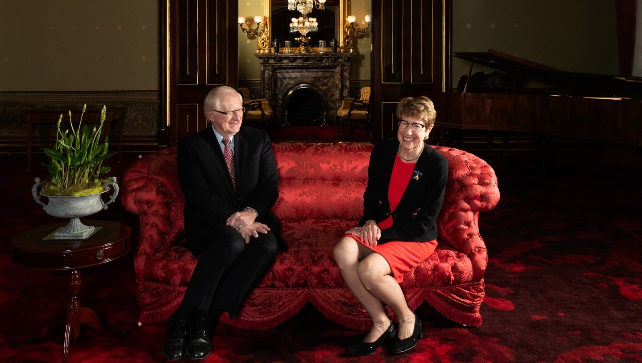 The vice regal couple seated on a plush, old style and bright red sofa. Mr Wilson, on the far left, is wearing a dark business suit. Governor Beazley on the far right, is in a black skirt with a red jacket. Both are smiling warmly. Behind them is a large entrance into another formal area and we can see a mirror reflecting chandeliers.