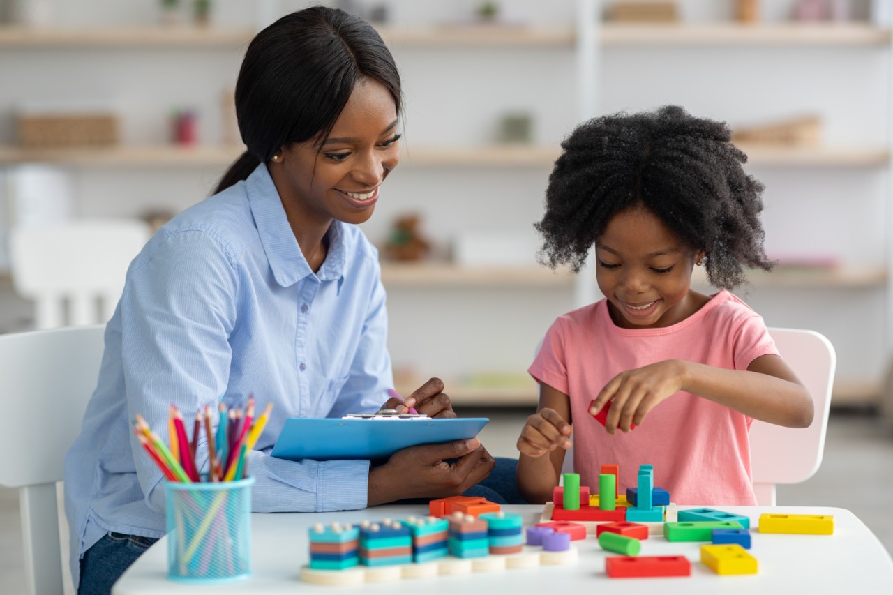 A young girl works through coloured play-based assessments with a smiling professional taking notes on a clipboard.