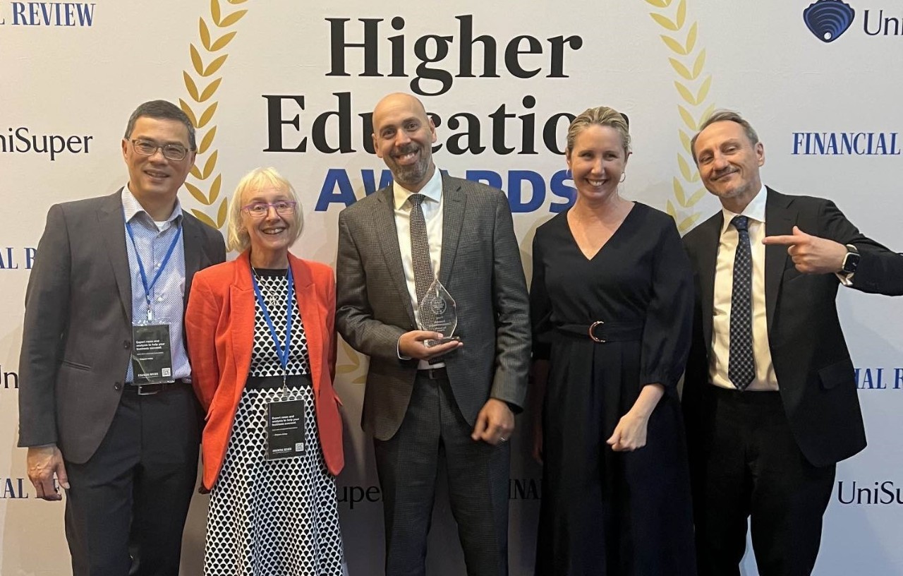 Image of a group of people standing together smiling, the person in the middle is holding a trophy.