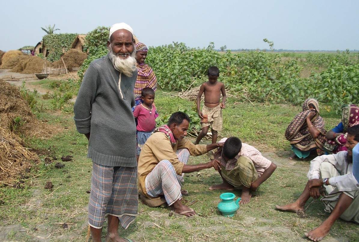 Under a clear, blue sky, several members of a rural Bangladeshi family are gathered. Standing in the foreground is a white-bearded elder looking at the camera.